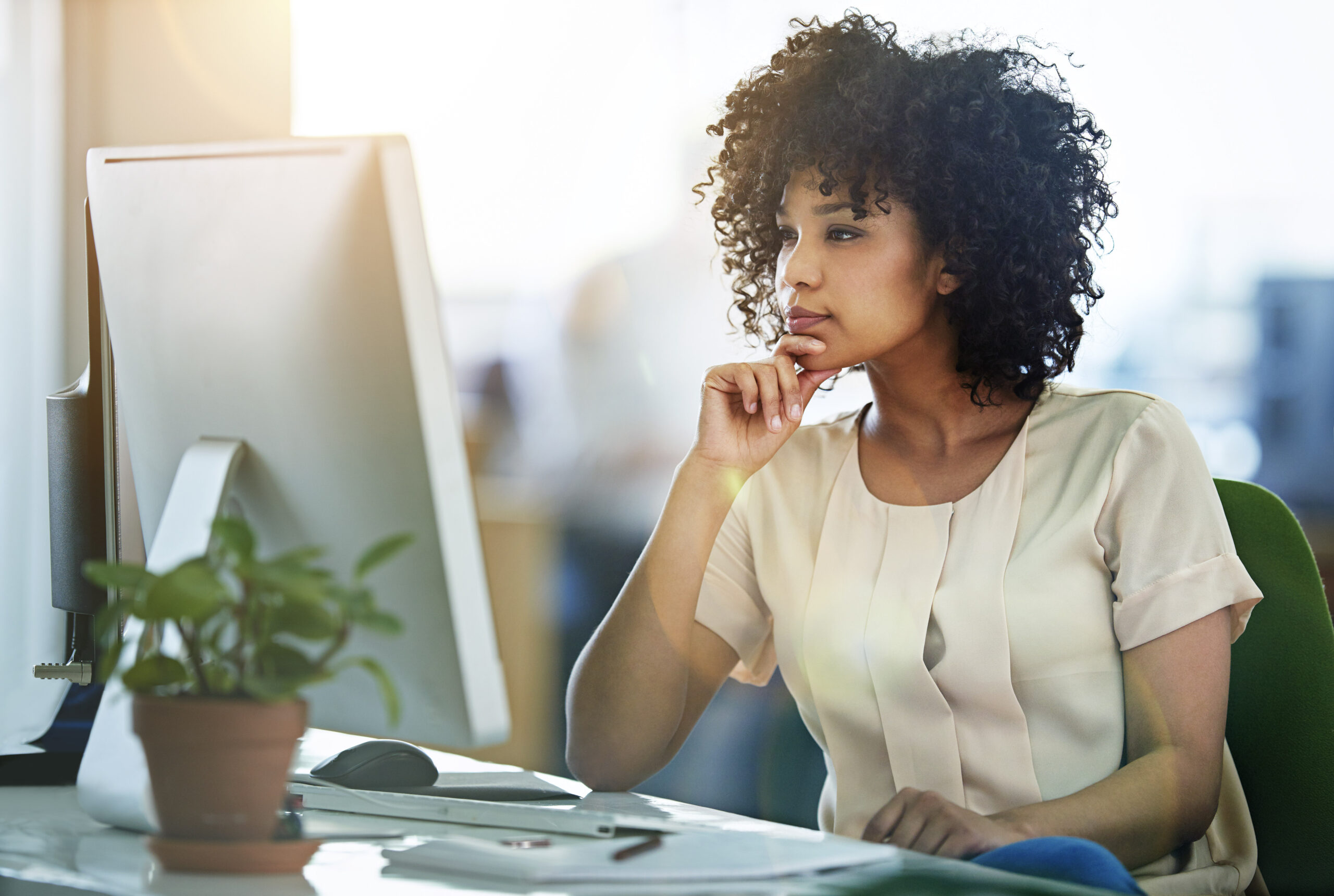 Woman wearing a beige blouse sitting in front of a monitor, thinking, small plant on the desk
