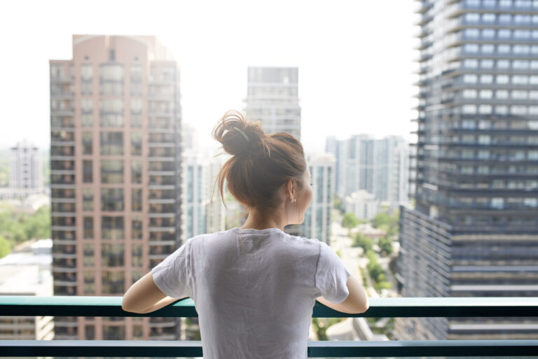 Young woman with her hair in a bun, standing on the balcony of her condominium, looking out at the city