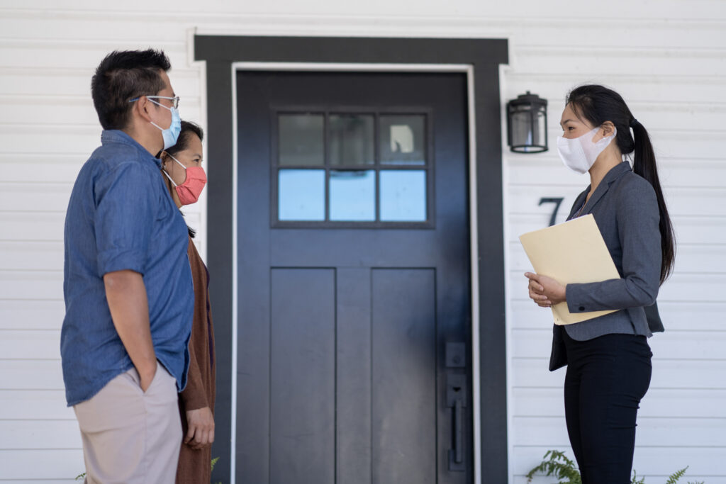Female realtor standing socially distanced from middle-aged couple clients outside a house, all wearing masks