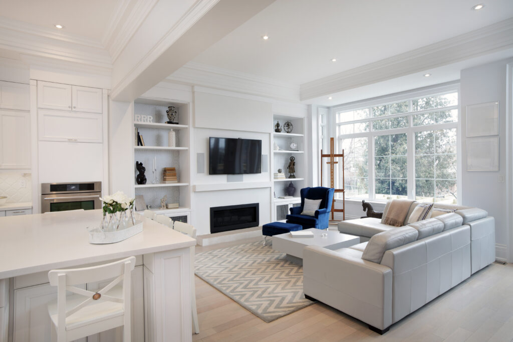 Modern, clean living room and kitchen with white couches, navy blue accent chair, white cabinets, television mounted on the wall