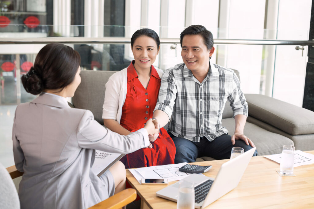 Asian couple, woman in red dress, sitting on a couch, smiling and shaking hands with a realtor