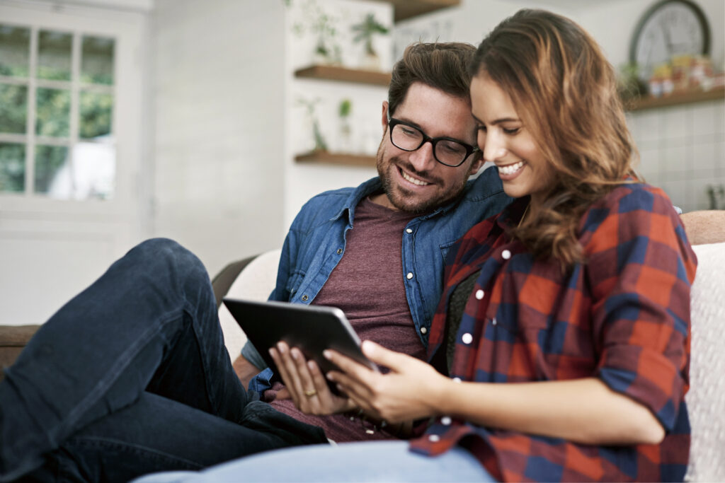 Young couple smiling, sitting on the couch reading blog post on a tablet, man in blue shirt, woman in plaid shirt