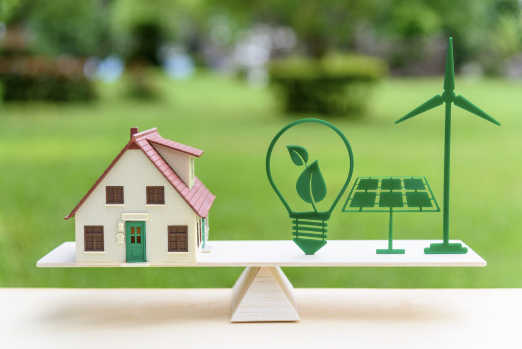 Toy house on a see-saw balanced by toy eco-light bulb, solar panel and windmill