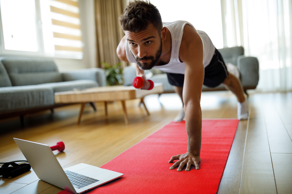 Athletic man in white tank, black shorts holding one-arm plank on a yoga mat in his living room; weighted row with other arm