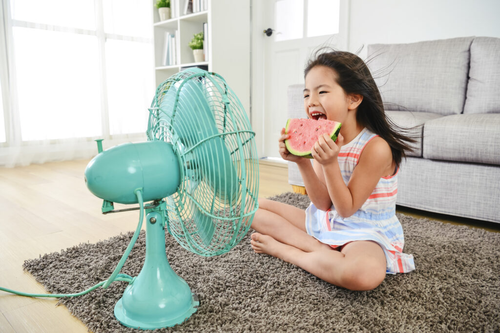 Young girl gleefully eating watermelon, seated in front of a blue fan, cooling herself on a hot summer day