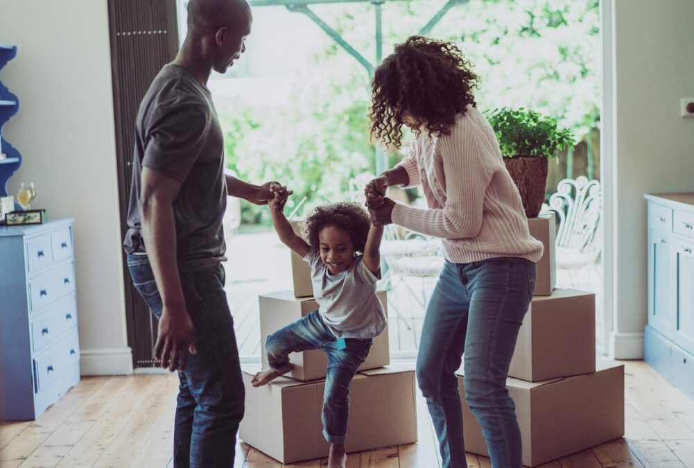 Parents playing with young child in their living room, with moving boxes and large window to yard in the background