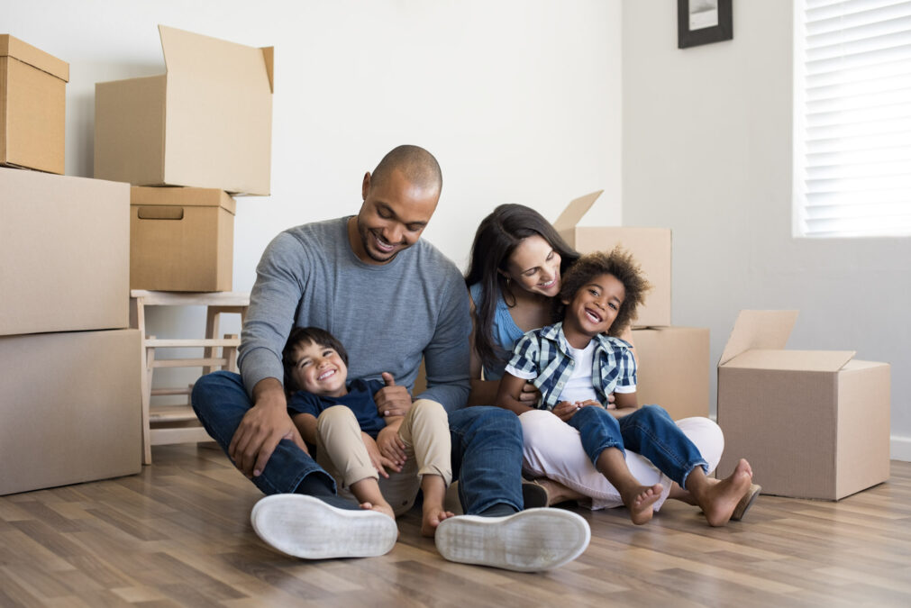A young couple with two sons sit on the floor in front of moving boxes