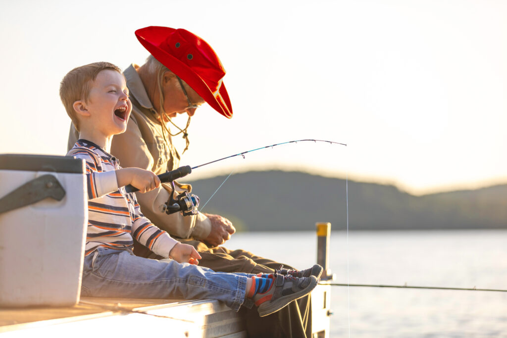 Young boy sits on a dock with his grandfather at sunset, fishing and laughing