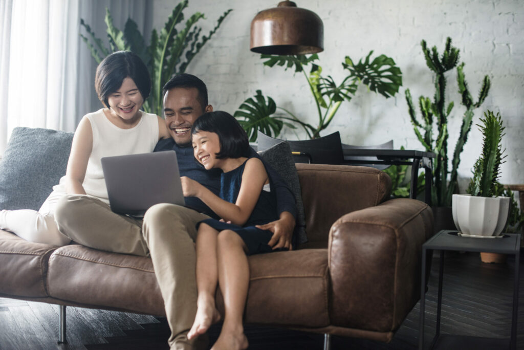 Happy couple sits on a couch with their young daughter looking at a laptop in a room with several plants