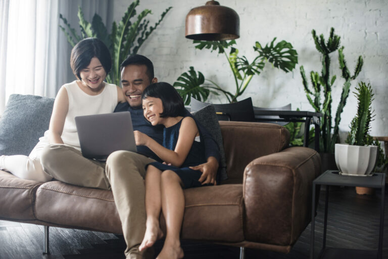 Happy couple sits on a couch with their young daughter looking at a laptop in a room with several plants