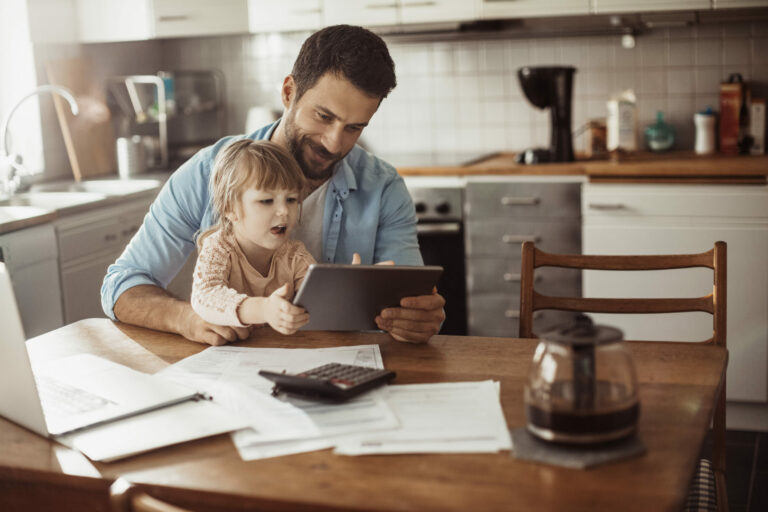 Young girl sitting on father’s lap in the kitchen while is he is working remotely