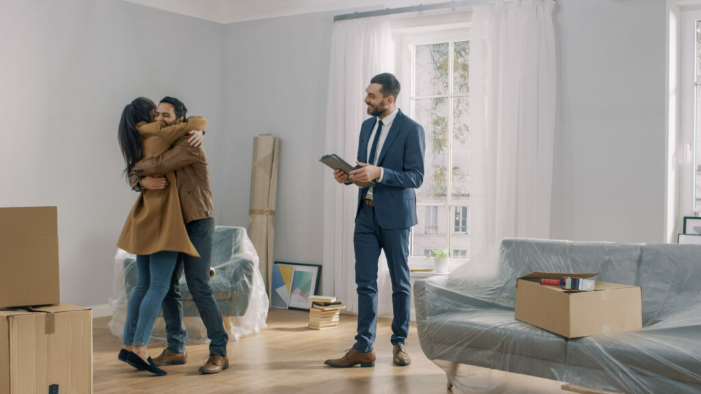 Young couple embracing ecstatically in their new home while real estate agent smiles
