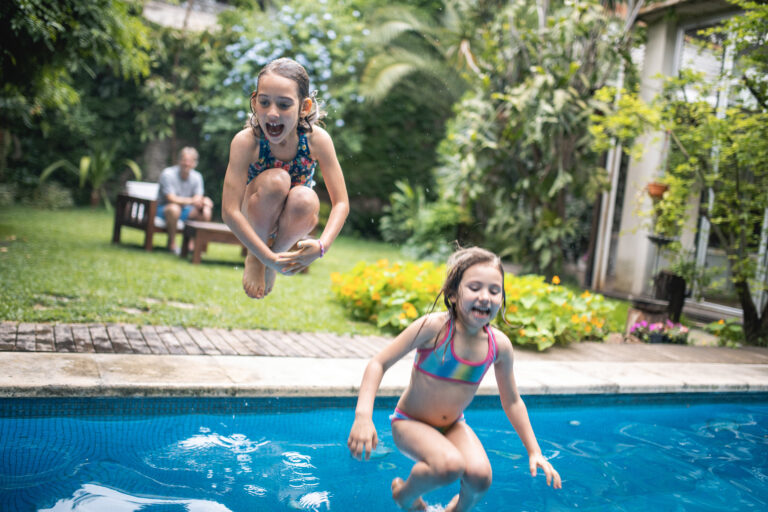 Two young kids jumping into a backyard pool, smiling and laughing, having fun