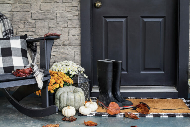 Close-up shot of exterior front door, black, accentuated with rocking chair, black and white plaid pillow and throw, pumpkins and gourds, orange flowers and black rain boots