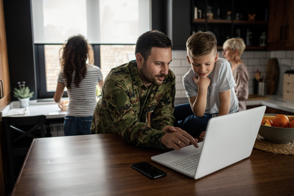 A man in military uniform sits at the kitchen counter with a laptop and young son
