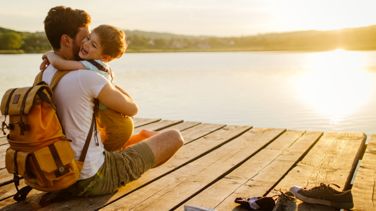 A man embraces a young boy at sunset on a dock overlooking the water