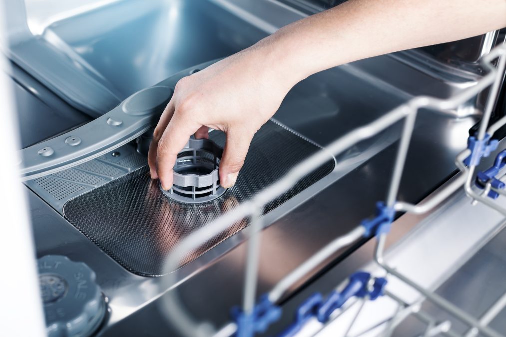 A hand reaches into the base of a dishwasher to change the filter
