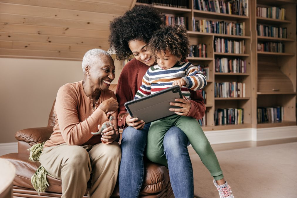 A grandmother, mother and child sit together reading a tablet in a library