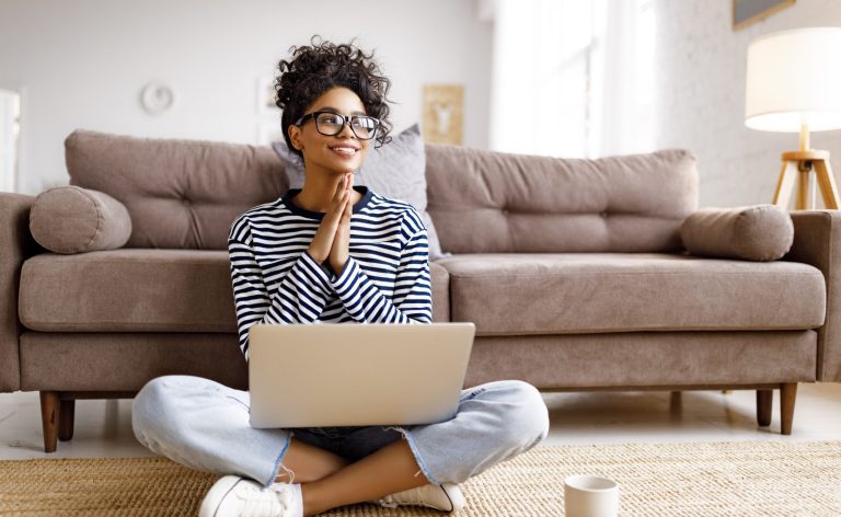 A young woman in a striped shirt and glasses sits in front of sofa with laptop