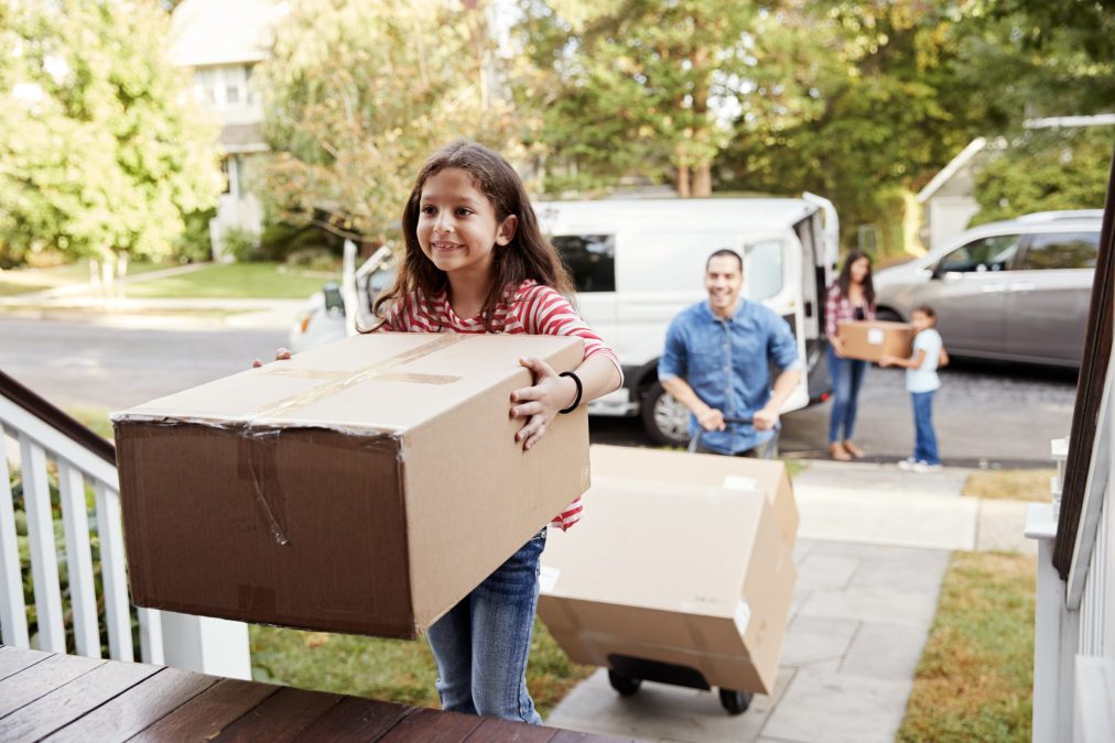 A young girl carries a moving box up the front steps of a home