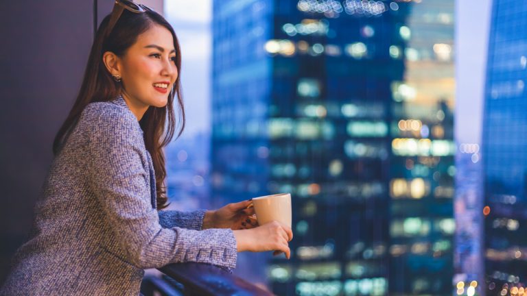 Woman enjoying the evening view from her balcony while looking at the urban skyscraper cityscape at night with a cup of hot coffee