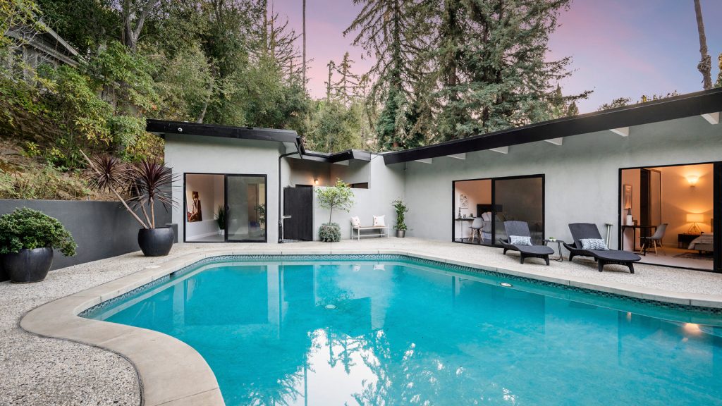 An inground pool and a white modern home surrounded by tall trees