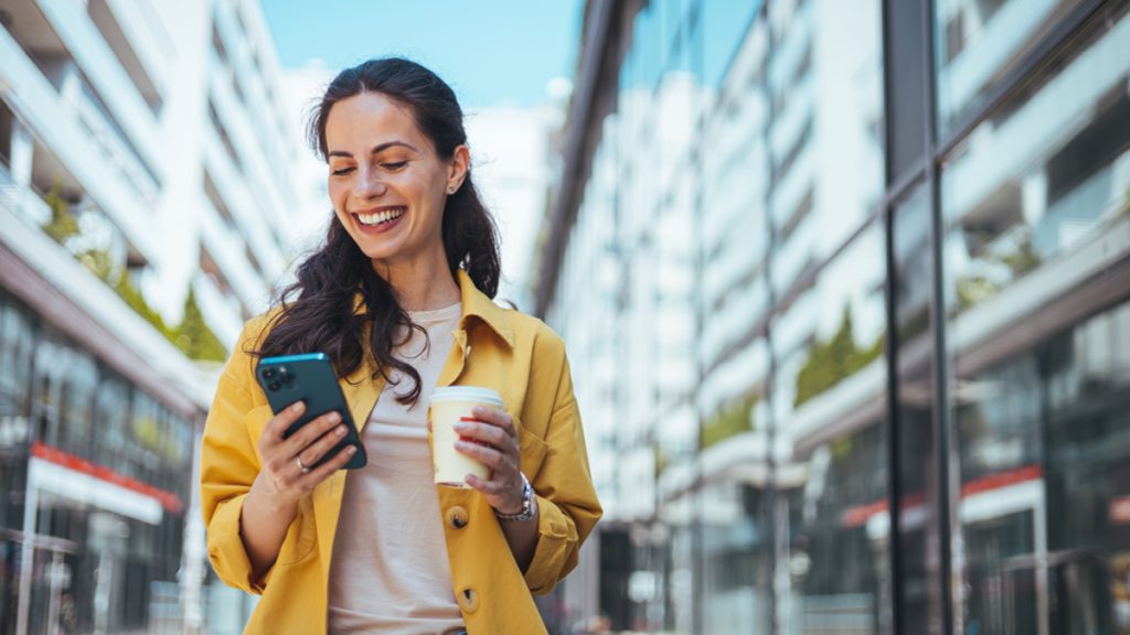 Woman in yellow coat surfing the net on smartphone while drinking coffee outdoors