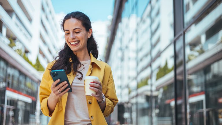 Woman in yellow coat surfing the net on smartphone while drinking coffee outdoors