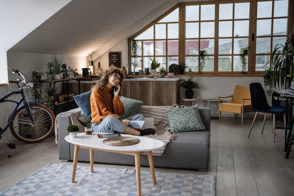 A young woman sitting on a grey sofa in an apartment talking on the phone