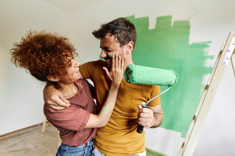 Cheerful couple having fun during home renovation process while woman is putting green paint on man’s face