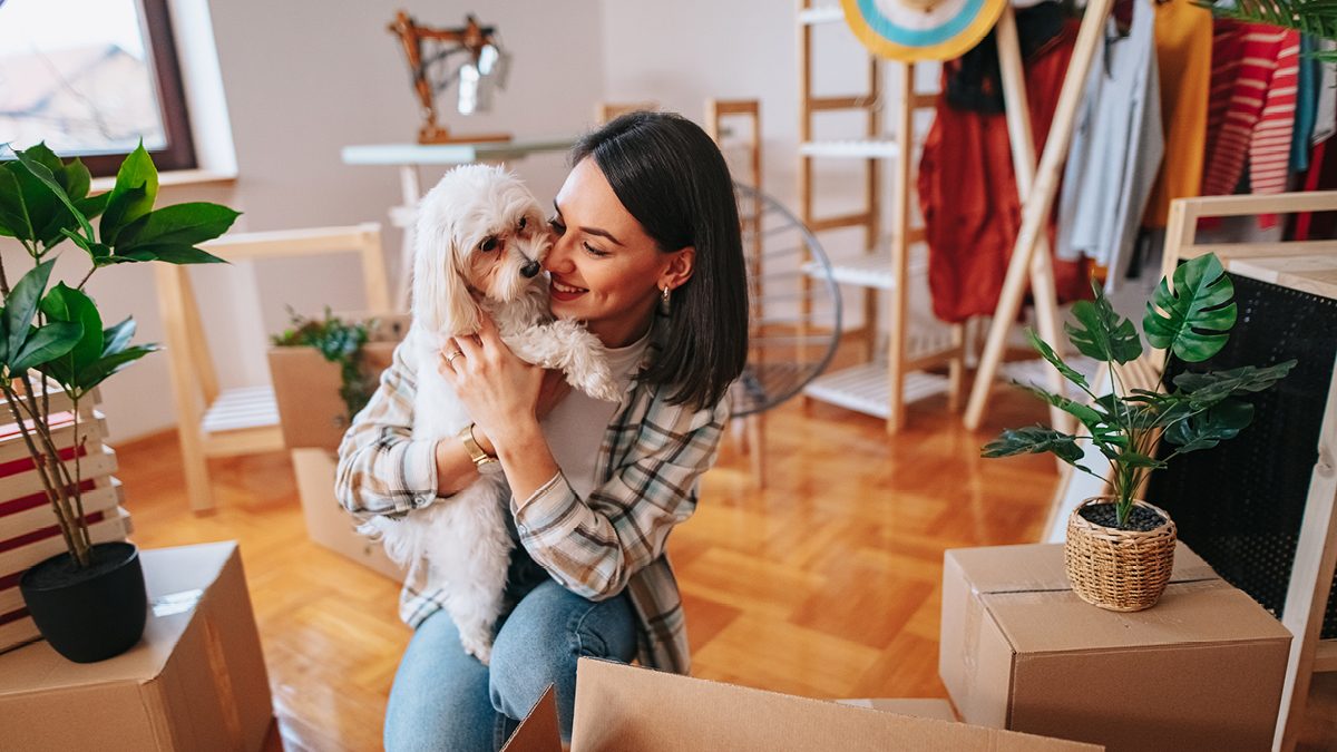 A young woman holding a small white dog surrounded by moving boxes
