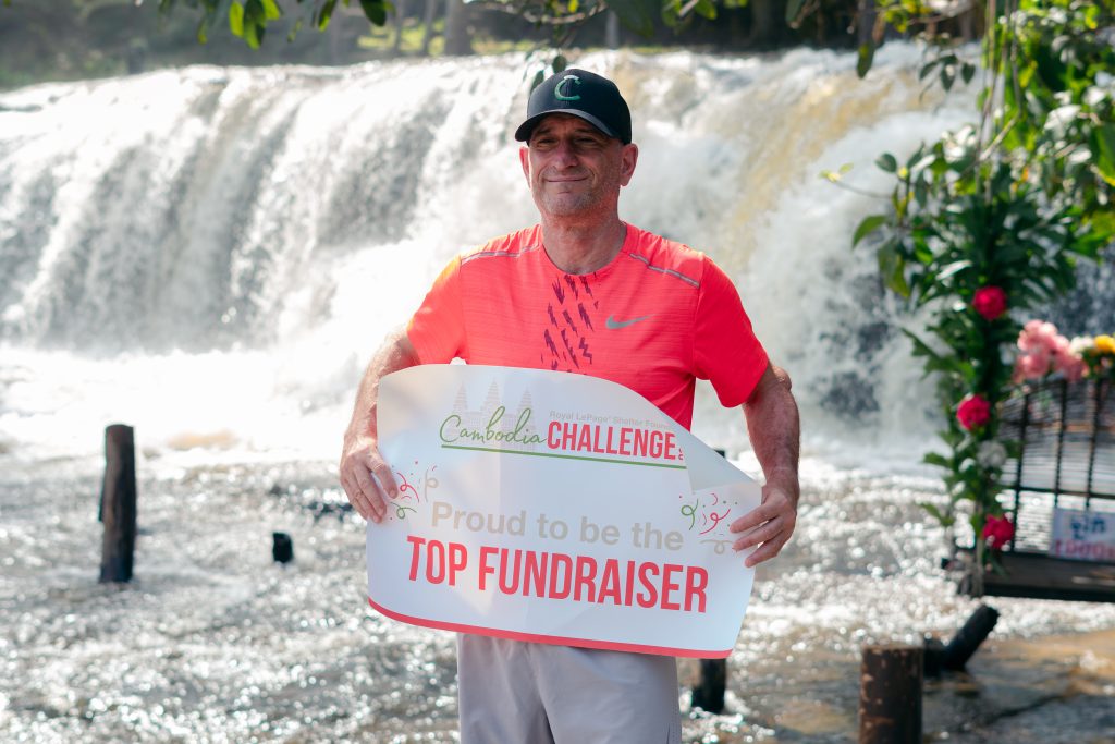A man in a red shirt holds a sign in front of a waterfall