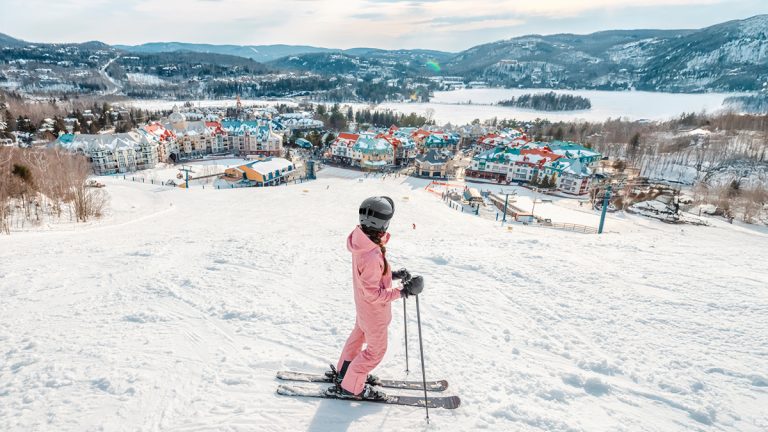 Skier at the top of a ski hill in a pink snowsuit looks out at the town colourful town below