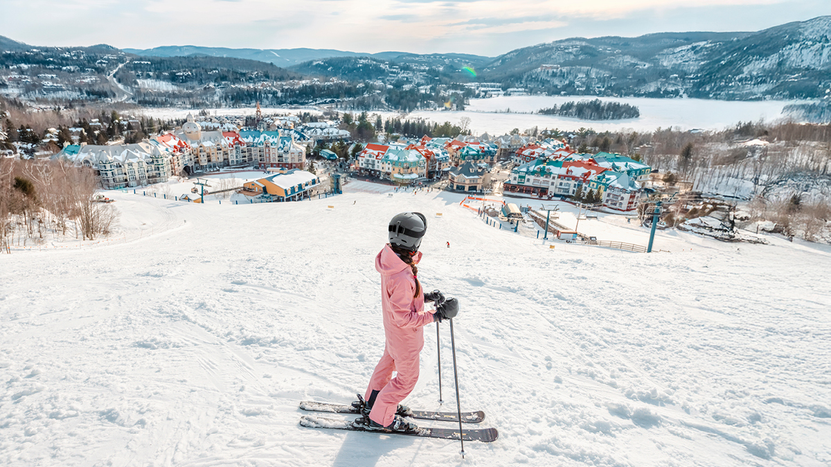 Skier at the top of a ski hill in a pink snowsuit looks out at the town colourful town below