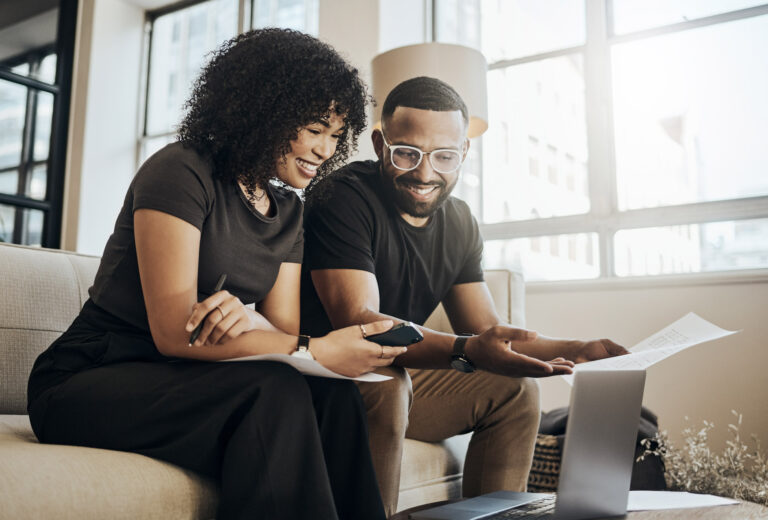 A young couple sit on a couch in a modern living room looking over finances on a laptop