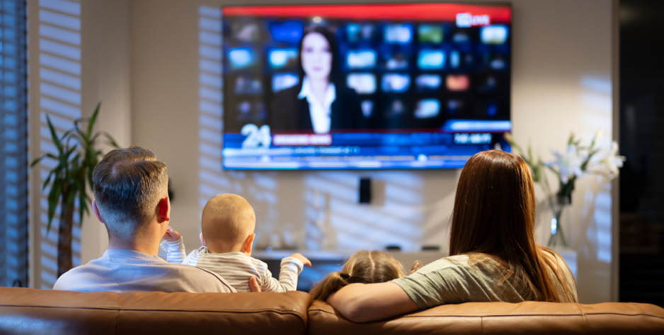 Family relaxing on leather couch, watching wall-mounted news broadcast in warmly lit living room with decorative plants.