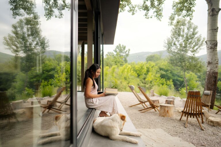 Photo of a young woman reading a book on the front porch of her cabin home, accompanied by her dog