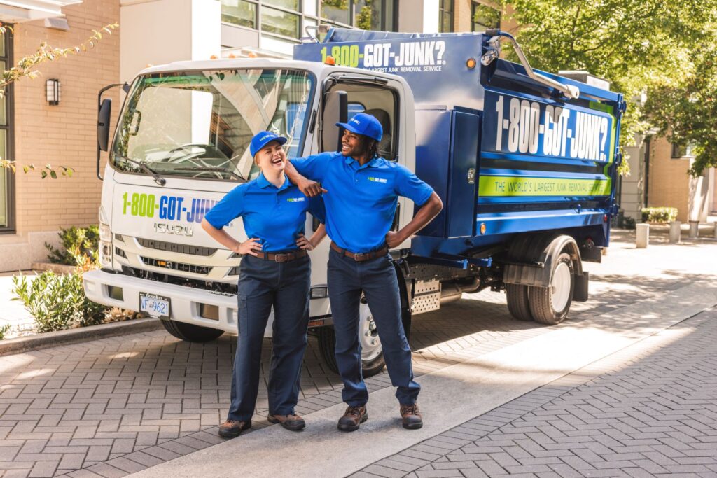 Two smiling 1-800-GOT-JUNK? team members in blue uniforms standing in front of a branded junk removal truck.
