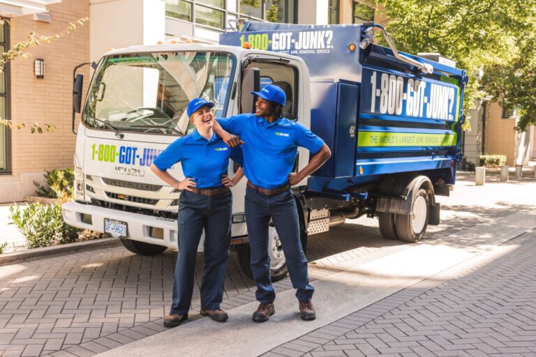 Two smiling 1-800-GOT-JUNK? team members in blue uniforms standing in front of a branded junk removal truck.